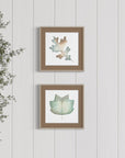 Two framed leaf prints on a white paneled wall with a plant to the left.