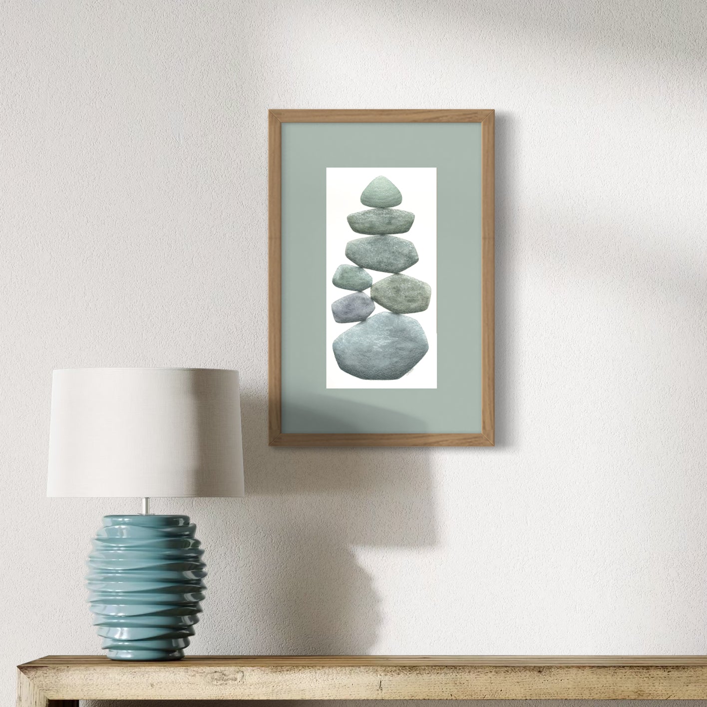 Framed artwork of stacked stones on a wall above a table with a lamp.