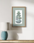 Framed artwork of stacked stones on a wall above a table with a lamp.