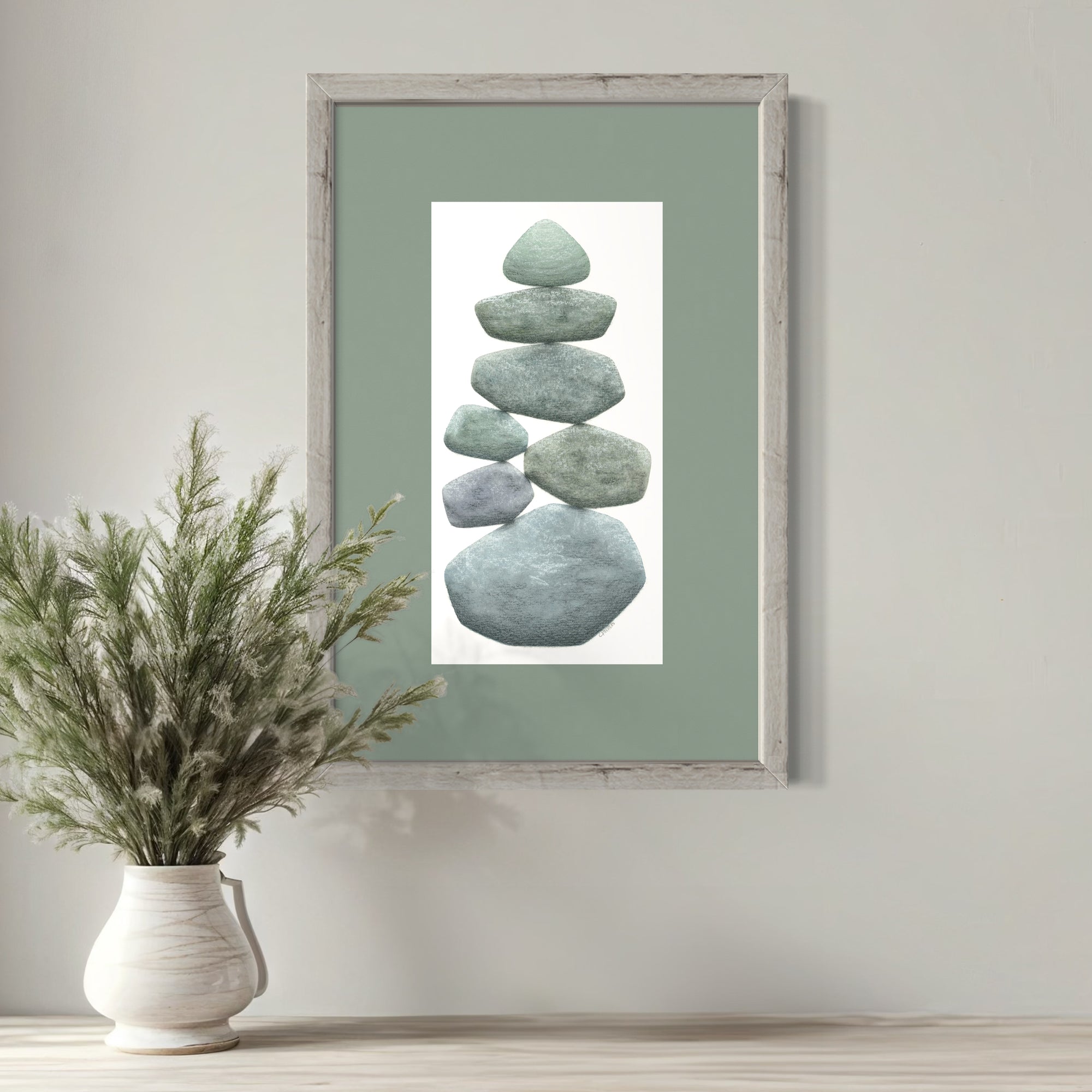 Framed artwork of stacked stones on a green background, placed above a vase with plants on a wooden surface.