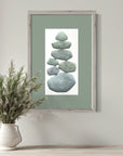 Framed artwork of stacked stones on a green background, placed above a vase with plants on a wooden surface.